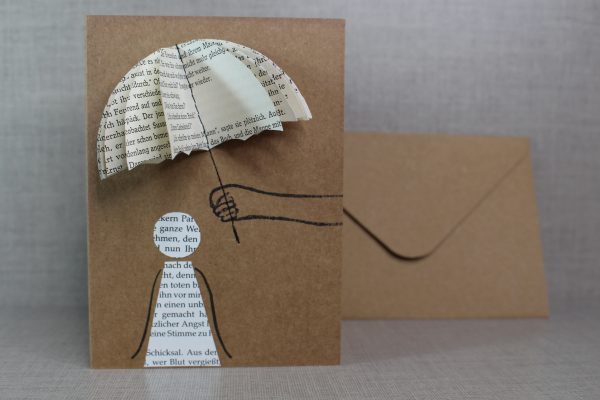 upcycling Grußgarte mit Buchseiten, Mensch sitzt am Rand, ein anderer hält den offenen Regenschirm